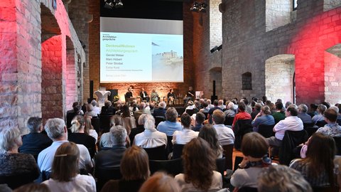 "Konferenzvortrag in einem historischen Gewölbesaal mit roten Backsteinmauern. Auf der Bühne sitzen etwa sieben Diskussionsteilnehmer. Ein großer Projektorschirm zeigt die Präsentation 'DenkmalWohnen - Architekturgespräche' mit Namen und Foto eines Schlosses. Das Publikum sitzt in Stuhlreihen und schaut zur Bühne. Die Wände sind mit rotem und gelbem Licht beleuchtet. Im Hintergrund massive Steinpfeiler."