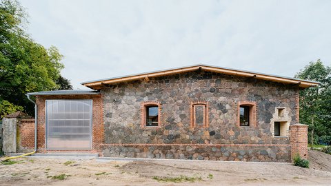 „Giebelseite eines sanierten Gebäudes aus Feldsteinen und rotem Backstein. Die Fassade hat mehrere schmale Fenster und links ein großes Tor aus lichtdurchlässigem Material.“