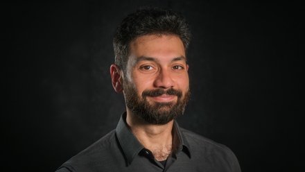 Porträt eines lächelnden, erwachsenen Mannes mit dunklen Haaren und Vollbart, Schulteransicht, er trägt ein dunkles Hemd; Kopf-Schulter-Aufnahme vor einem dunklen, unscharfen Hintergrund.