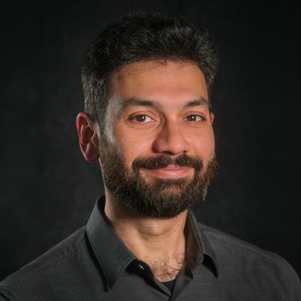 Porträt eines lächelnden, erwachsenen Mannes mit dunklen Haaren und Vollbart, Schulteransicht, er trägt ein dunkles Hemd; Kopf-Schulter-Aufnahme vor einem dunklen, unscharfen Hintergrund.