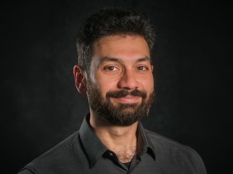 Porträt eines lächelnden, erwachsenen Mannes mit dunklen Haaren und Vollbart, Schulteransicht, er trägt ein dunkles Hemd; Kopf-Schulter-Aufnahme vor einem dunklen, unscharfen Hintergrund.