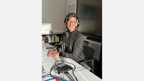 Angelika Hinterbrandner sitzt lächelnd an einem Schreibtisch vor einem professionellen Mikrofon. Sie trägt Kopfhörer und ist bereit für eine Podcast-Aufnahme; im Vordergrund ist ein mobiles Aufnahmegerät zu sehen.