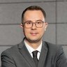 „Porträt eines Mannes mit Brille im dunkelgrauen Anzug, der mit verschränkten Armen in einem modernen Bürogebäude steht und in die Kamera blickt.“