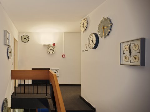 "Die Ansicht eines Flurs und Treppenhauses in einem Gebäude. Die Wände sind weiß gestrichen. An allen Wänden hängen zahlreiche, unterschiedliche Wanduhren in verschiedenen Größen und Stilen, von schlichten runden Uhren bis hin zu quadratischen und Uhren mit Zahnradmotiven. Die Treppe hat einen hellen Holzhandlauf. Am Ende des Flurs ist ein roter Feuermelder zu sehen."
