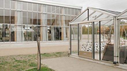 Verglaste Hallenfassade mit davorstehendem kleinem Gewächshaus‑Pavillon; Kiesweg und Rasen im Vordergrund, bewölkter Himmel.