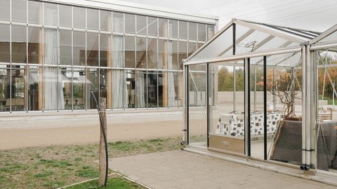 Verglaste Hallenfassade mit davorstehendem kleinem Gewächshaus‑Pavillon; Kiesweg und Rasen im Vordergrund, bewölkter Himmel.