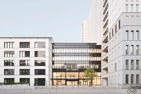 Moderne Stadtarchitektur mit drei mehrstöckigen Gebäuden um einen zentralen Platz. Im Zentrum ein Gebäude mit großer Glasfassade im Erdgeschoss, beschriftet mit 'BIH CHARITÉ' und 'RAHM HIRSCH CENTER'. Die umliegenden Gebäude sind weiß verputzt mit regelmäßig angeordneten Fenstergruppen. Rechts zwei Fahrräder an einer Parkanlage, am Boden gepflasterte Platzfläche mit Metallbügeln. Klarer blauer Himmel.