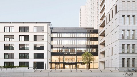 Moderne Stadtarchitektur mit drei mehrstöckigen Gebäuden um einen zentralen Platz. Im Zentrum ein Gebäude mit großer Glasfassade im Erdgeschoss, beschriftet mit 'BIH CHARITÉ' und 'RAHM HIRSCH CENTER'. Die umliegenden Gebäude sind weiß verputzt mit regelmäßig angeordneten Fenstergruppen. Rechts zwei Fahrräder an einer Parkanlage, am Boden gepflasterte Platzfläche mit Metallbügeln. Klarer blauer Himmel.
