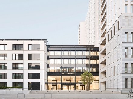 Moderne Stadtarchitektur mit drei mehrstöckigen Gebäuden um einen zentralen Platz. Im Zentrum ein Gebäude mit großer Glasfassade im Erdgeschoss, beschriftet mit 'BIH CHARITÉ' und 'RAHM HIRSCH CENTER'. Die umliegenden Gebäude sind weiß verputzt mit regelmäßig angeordneten Fenstergruppen. Rechts zwei Fahrräder an einer Parkanlage, am Boden gepflasterte Platzfläche mit Metallbügeln. Klarer blauer Himmel.