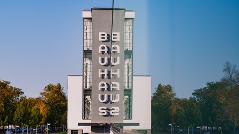 Fassade des weißen Bauhaus-Gebäudes mit vertikal angeordneten Buchstaben "BAUHAUS" und "WEIMAR" vor blauem Himmel und Bäumen.
