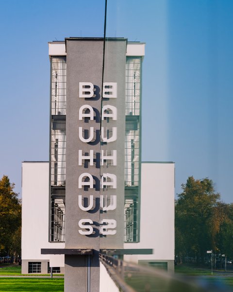 Fassade des weißen Bauhaus-Gebäudes mit vertikal angeordneten Buchstaben "BAUHAUS" und "WEIMAR" vor blauem Himmel und Bäumen.