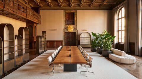 in heller Konferenzraum verbindet historische Architektur mit modernem Design. Ein langer Holztisch mit Stühlen steht unter einer reich verzierten Kassettendecke, daneben große Fenster, Pflanzen und hohe Regale.