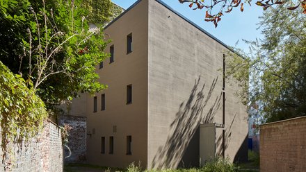 Ein kompakter, monolithisch wirkender Neubau mit ruhiger Fassadengestaltung steht in einem begrünten Hinterhof zwischen alten Mauern, die reduzierte Architektur tritt bewusst zurück und ordnet sich dem Ort unter, wodurch das Bild Nachverdichtung im Bestand zeigt und für respektvollen Umgang mit gewachsenen Strukturen und neue Wohnformen im innerstädtischen Kontext steht.