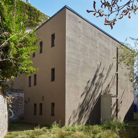 Ein kompakter, monolithisch wirkender Neubau mit ruhiger Fassadengestaltung steht in einem begrünten Hinterhof zwischen alten Mauern, die reduzierte Architektur tritt bewusst zurück und ordnet sich dem Ort unter, wodurch das Bild Nachverdichtung im Bestand zeigt und für respektvollen Umgang mit gewachsenen Strukturen und neue Wohnformen im innerstädtischen Kontext steht.