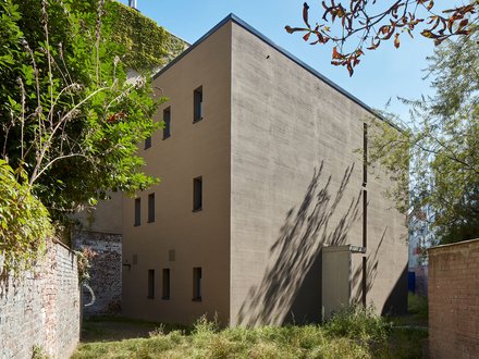 Ein kompakter, monolithisch wirkender Neubau mit ruhiger Fassadengestaltung steht in einem begrünten Hinterhof zwischen alten Mauern, die reduzierte Architektur tritt bewusst zurück und ordnet sich dem Ort unter, wodurch das Bild Nachverdichtung im Bestand zeigt und für respektvollen Umgang mit gewachsenen Strukturen und neue Wohnformen im innerstädtischen Kontext steht.