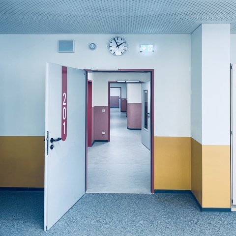 "Großformatiger Flur mit hellblauer Decke und punktförmigem Muster. Wände oben weiß, unten gelb. Eine offene Tür mit dunkelrotem Rahmen zeigt die Nummer '201' und führt in einen Innenraum mit dunkelblauen Türrahmen. Weitere Türen und Raumteilwände sichtbar. Eine Wanduhr mit schwarzem Rand und schwarzen Zeigern zeigt die Zeit an."
