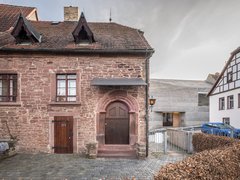 "Historisches Steingebäude mit rotem Ziegeldach und zwei Dachgauben. Die Fassade besteht aus rötlichem Sandstein mit zwei symmetrischen Fensteröffnungen und zwei Eingangstüren. Der Haupteingang hat einen rundbogigen Rahmen in Terrakotta-Farbe. Rechts daneben ein modernes weißes Gebäude mit Fachwerk. Davor ein gepflasterter Platz mit einer Metallbarriere und einer grünen Hecke. Der Himmel ist bewölkt."