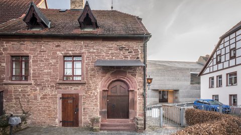 "Historisches Steingebäude mit rotem Ziegeldach und zwei Dachgauben. Die Fassade besteht aus rötlichem Sandstein mit zwei symmetrischen Fensteröffnungen und zwei Eingangstüren. Der Haupteingang hat einen rundbogigen Rahmen in Terrakotta-Farbe. Rechts daneben ein modernes weißes Gebäude mit Fachwerk. Davor ein gepflasterter Platz mit einer Metallbarriere und einer grünen Hecke. Der Himmel ist bewölkt."