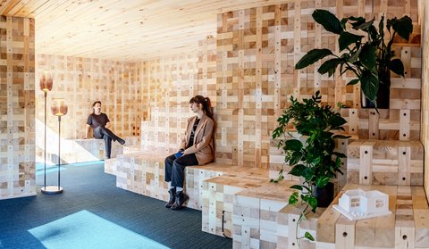 Zwei Personen sitzen auf gestuften Holzbänken in einem modularen Holzraum mit Pflanzen.