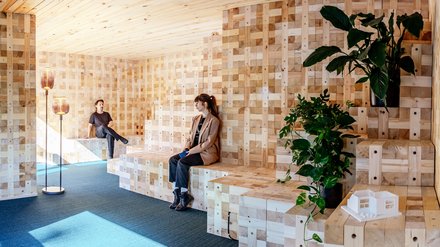 Zwei Personen sitzen auf gestuften Holzbänken in einem modularen Holzraum mit Pflanzen.
