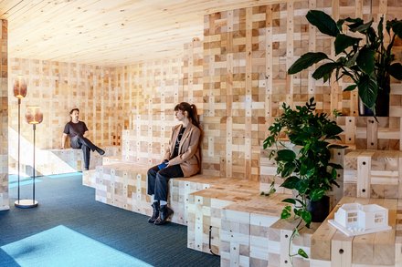 Zwei Personen sitzen auf gestuften Holzbänken in einem modularen Holzraum mit Pflanzen.