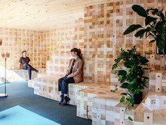 Zwei Personen sitzen auf gestuften Holzbänken in einem modularen Holzraum mit Pflanzen.