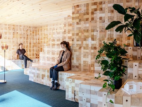 Zwei Personen sitzen auf gestuften Holzbänken in einem modularen Holzraum mit Pflanzen.