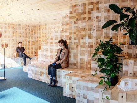 Zwei Personen sitzen auf gestuften Holzbänken in einem modularen Holzraum mit Pflanzen.