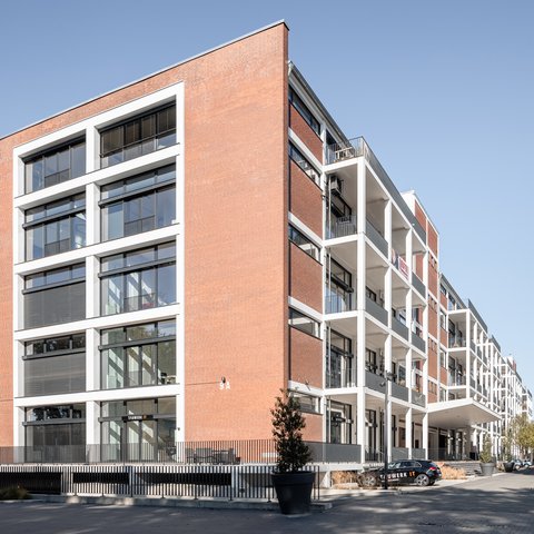 Mehrstöckiges Gebäude mit roter Backsteinfassade und großen Fenstern an einer Straße mit parkenden Autos