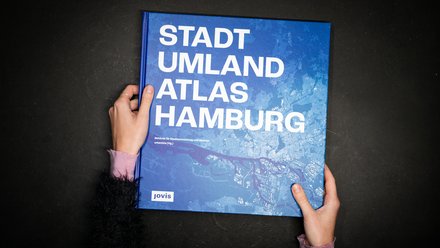 Hände halten das Buch „Stadt Umland Atlas Hamburg“ vor dunklem Hintergrund.