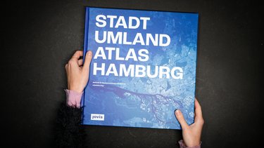 Hände halten das Buch „Stadt Umland Atlas Hamburg“ vor dunklem Hintergrund.