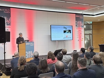 Vortrag bei der BIM World Munich: Person am Rednerpult spricht, Publikum sitzt zugewandt. Rechts eine große Leinwand mit Folie zum Projekt „Sanierung und Umnutzung des Jugendstilparks Haar“.
