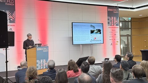 Vortrag bei der BIM World Munich: Person am Rednerpult spricht, Publikum sitzt zugewandt. Rechts eine große Leinwand mit Folie zum Projekt „Sanierung und Umnutzung des Jugendstilparks Haar“.