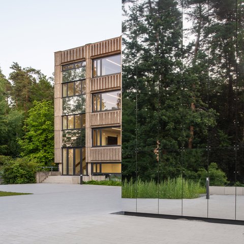 Spiegelnde Glasfassade eines modernen Gebäudes neben einem mehrstöckigen Gebäude mit großen Fenstern und umgeben von Bäumen