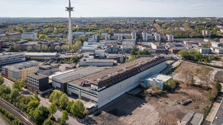 Luftaufnahme eines Stadtviertels. Im Vordergrund ein langes, graues Industriegebäude; rechts daneben ein brachliegendes, erdiges Areal mit wenigen Bäumen. Links Straße und Bahngleise. In der Mitte ragt ein Funkturm auf, dahinter dichte Wohn- und Bürohäuser unter blauem Himmel.