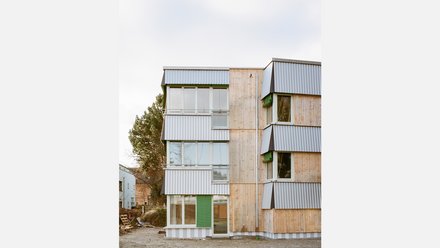 Wohngebäude mit Holz- und Wellblechfassade; versetzte Fensterbänder und grüne Rollläden; Erdgeschoss im Ausbau.