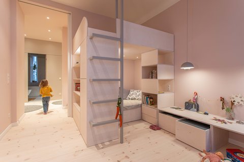 Helles Kinderzimmer mit Hochbett, Leiter, Schreibtisch und spielendem Kind im Hintergrund.