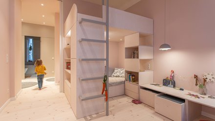 Helles Kinderzimmer mit Hochbett, Leiter, Schreibtisch und spielendem Kind im Hintergrund.