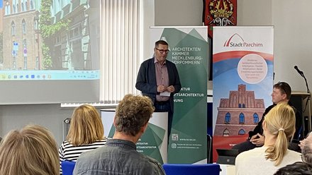 Redner begrüßt das Publikum vor Leinwand und Veranstaltungsbannern in einem Saal.