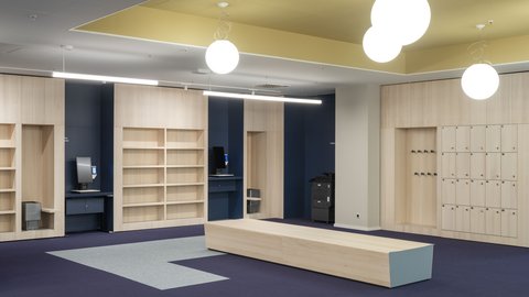 "Großzügiges modernes Bibliotheksfoyer. Gelbe Deckenzone mit weißen Kugelleuchten. Beidseitig Holzregale in Natur- und Dunkelblau mit offenen Fächern. Zentral ein helles Holz-Sitzelement. Dunkelblaue Bodenzone mit hellgrauem Teppichstreifen. Links ein Serviceschalter mit Monitor. Rechts weiße Wandelemente und blaue Türen. Moderne Spot-Beleuchtung."