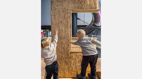 "Zwei Kinder erkunden eine Ausstellung mit einer Wand aus Stroh. Eines steht auf einem kleinen Podest und greift nach einem Holzfenster, das in die Wand eingebaut ist, während das andere die Oberfläche mit der Hand berührt. Die Szene vermittelt spielerisches Entdecken von Materialien."