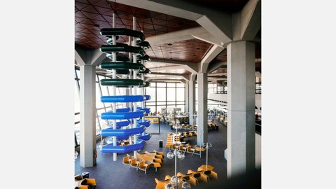 Farbfoto aus der Vogelperspektive auf eine modernistische Wartehalle mit Betonstützen und rötlicher Rautendecke. Links hängt eine markante Skulptur aus gestapelten dunkelgrünen und blauen Rohrringen. Am Boden sind Gruppen von leuchtend gelben Sitzschalen angeordnet. Im Hintergrund befindet sich ein Bar-Bereich mit "SNACK"-Schild. Große Fensterfronten lassen viel Tageslicht herein.