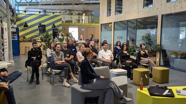 Publikum hört einem Vortrag in einem hellen, industriell geprägten Innenraum zu.