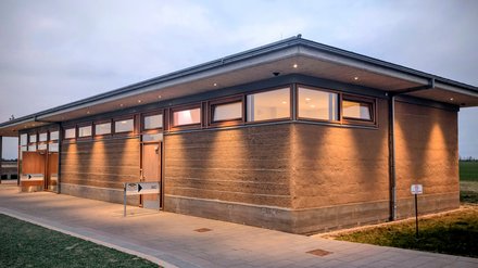 Dämmerung: Besucherzentrum mit Vordach und Oberlichtfenstern, warm beleuchtet; gepflasterter Weg, Wegweiser „WC“.