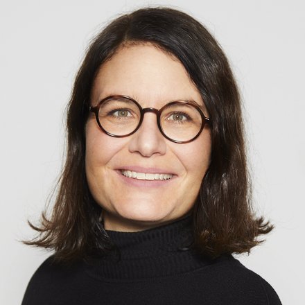 Porträtfoto: Person mit runder Brille, schulterlangem dunklem Haar und schwarzem Rollkragen, lächelnd vor hellem Hintergrund.