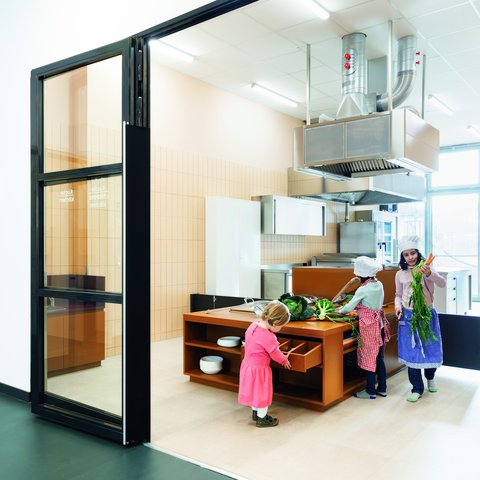"Offene Lernküche hinter schwarzen Rahmenschiebetüren. Innenraum mit beige gefliesten Wänden. Orange-braunes Küchenblock-Element mit integrierten Arbeitsplatten und Schränken. Metall-Dunstabzugshaube an der Wand. Drei Kinder in Kostümen beschäftigen sich mit Gemüse und Kochutensilien. Heller Bodenbelag, natürliche und künstliche Beleuchtung."