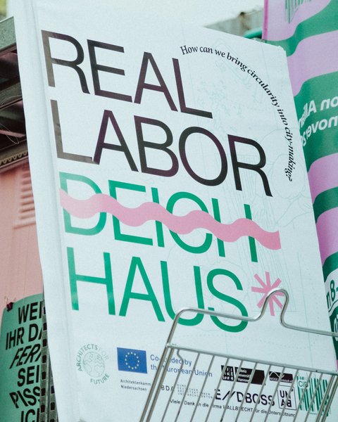 Großformatiges Plakat mit der Aufschrift „REAL LABOR DEICH HAUS“ in Schwarz und Grün sowie pinken Gestaltungselementen. Das Plakat steht in einem Ausstellungsbereich und wird von weiteren grün-pink gestalteten Informationsflächen umgeben.
