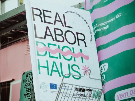 Großformatiges Plakat mit der Aufschrift „REAL LABOR DEICH HAUS“ in Schwarz und Grün sowie pinken Gestaltungselementen. Das Plakat steht in einem Ausstellungsbereich und wird von weiteren grün-pink gestalteten Informationsflächen umgeben.