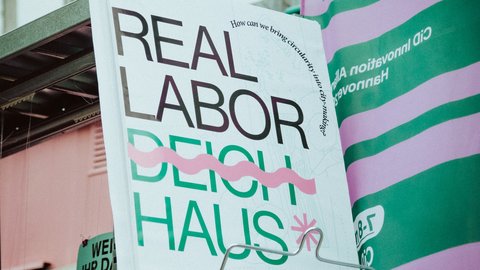Großformatiges Plakat mit der Aufschrift „REAL LABOR DEICH HAUS“ in Schwarz und Grün sowie pinken Gestaltungselementen. Das Plakat steht in einem Ausstellungsbereich und wird von weiteren grün-pink gestalteten Informationsflächen umgeben.