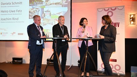 Podiumsgespräch mit vier Personen auf einer Bühne vor Präsentationsfolie.